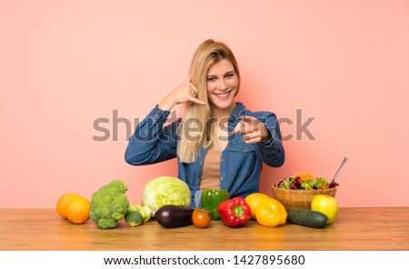Young blonde woman with many vegetables making phone gesture and pointing front