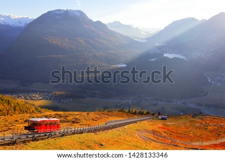 A funicular train travels on the hillside of meadows in orange color at sunset, overlooking Pontresina & St Motritz villages & lakes in Engadin Valley under alpine mountains in Grisons, Switzerland Royalty-Free Stock Photo #1428133346