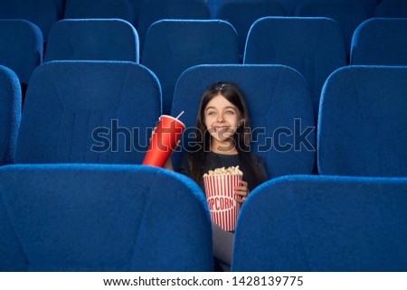 Front view of cheerful girl keeping drink, eating popcorn and watching funny film in movie house. Beautiful child sitting alone in hall, looking at screen and laughing. Concept of cinema and fun.
