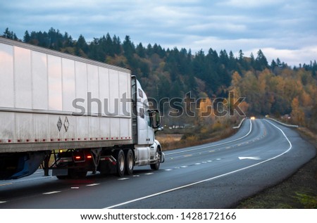 Big rig white long haul semi truck with semi trailer running with turned on headlights on rain dusted wet raining highway driving with another cars traffic in twilight evening time Royalty-Free Stock Photo #1428172166