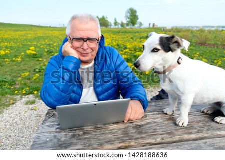 old age, technology, senior people, lifestyle, distance learning. Happy and Smiling Senior man 70-75 years old sitting in summer park with dog jack russell terrier and uses laptop computer.