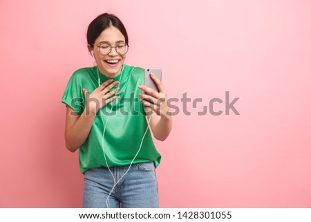 Portrait of a happy young casual girl standing isolated over pink background, having video call with earphones and mobile phone