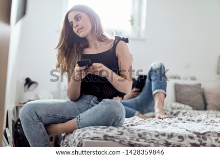 young brunette sitting on bed with her smart phone and her boyfriend with smart devide in the background, laying on bed as well