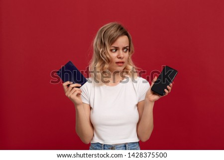 Portrait of a girl with curly blond hair in a white t-shirt on a red background. Displeased model looks at the screen of a mobile phone holding a passport.