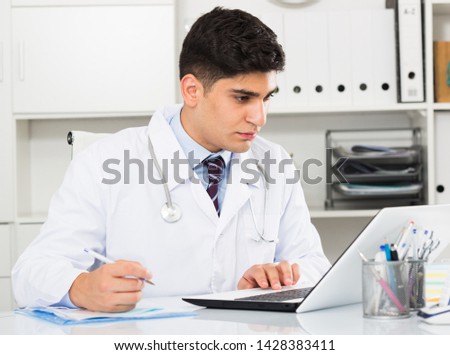 Male doctor 28-35 years old is doing report about patients in hospital. Royalty-Free Stock Photo #1428383411