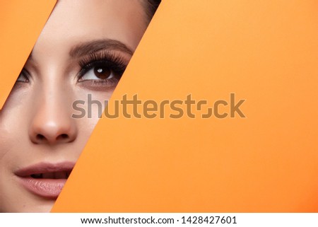 A girl with beautiful curry bright beautiful eyes with brown shadows and expressive eyebrows looks into the hole of colored paper.Fashion, beauty, make-up, cosmetics, make-up artist, beauty salon,busi