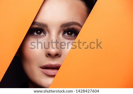 A girl with beautiful curry bright beautiful eyes with brown shadows and expressive eyebrows looks into the hole of colored paper.Fashion, beauty, make-up, cosmetics, make-up artist, beauty salon,busi
