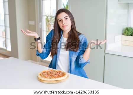 Beautiful young woman eating homemade tasty pizza at the kitchen clueless and confused expression with arms and hands raised. Doubt concept.