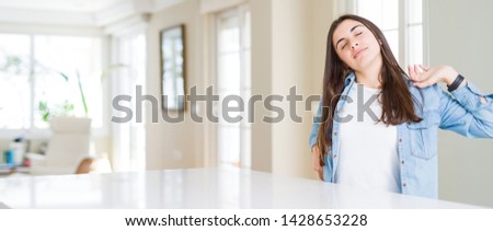 Wide angle picture of beautiful young woman sitting on white table at home stretching back, tired and relaxed, sleepy and yawning for early morning