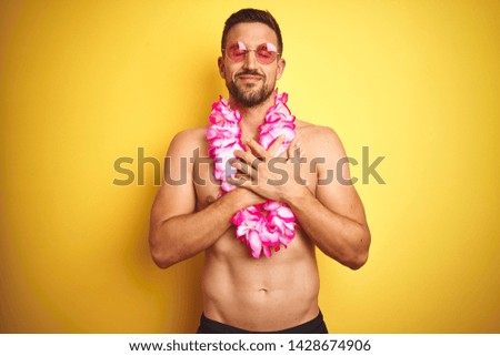 Young handsome shirtless man wearing sunglasses and pink hawaiian lei over yellow background smiling with hands on chest with closed eyes and grateful gesture on face. Health concept.