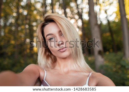 Young stylish blonde woman in a striped pink fashionable sundress makes selfie standing in the forest on a bright sunny summer day. Cute girl with blue eyes is taking a picture of herself.
