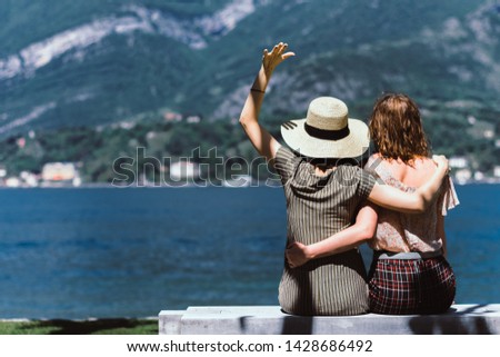 Lesbian couple, young women, girls, lovers sit at the lake, at the seaside on the bench looking at mountains and lake or sea with the picturesque view. 