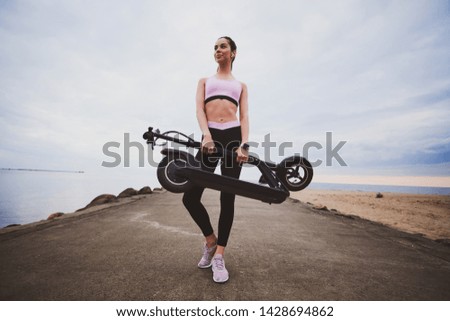 Low angle photo of pretty pensive girl with folded scooter in her hands. There are cloudy sky at background.