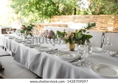 Wedding table setting decorated with fresh flowers in a brass vase. Wedding floristry. Banquet table for guests outdoors with a view of green nature. Bouquet with roses, eustoma and eucalyptus leaves.