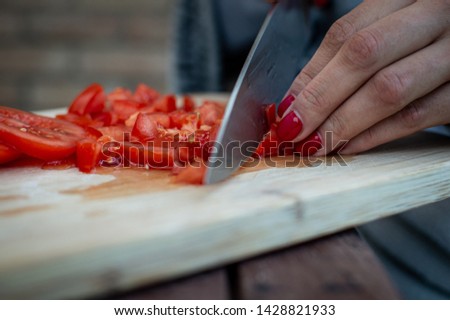 Picture of Woman's hands cutting tomato, behind fresh vegetables. Cuting board, light wood. Woman preparing food, with beautiful red nails and big knife.