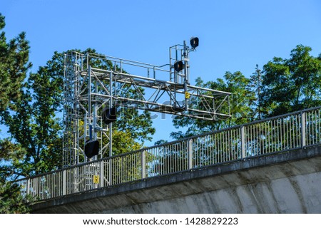 Railway signals and signs on metal frame among trees above bridge with fence.