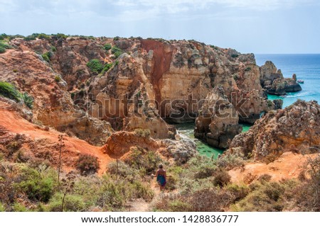 Landscape photo of a woman hiking down a hill with a view of the Portugal coast with big cliffs, sightseeing jet boats and turquoise waters. Shot in the Faro district  in the Algarve, Portugal. 