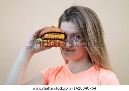 young attractive teen girl stands against an orange wall in an orange T-shirt and holds a big burger, she puts it to her face and looks into the camera