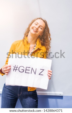 Young millennial teenage with red hair and a funny expression ,making lips and winking, holds a sign with the text " #genz" written on against a city wall and light flares
