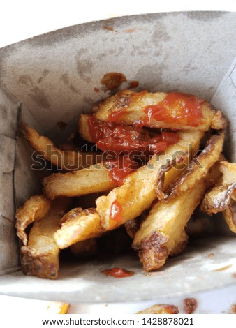 Few French fries with red ketchup in a greyish cardboard box with a glympse of some fries on a white table