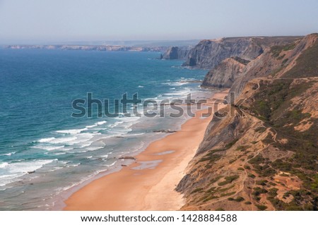 Aerial landscape photo of big, steep cliffs by the ocean with a huge, deserted beach at the bottom on a sunny afternoon. Shot in Praia da Cordoama near Sagres, Portugal. 