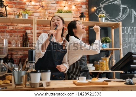 Two excited female colleagues in aprons working in cafe bar counter singing and dancing. relaxed woman waitresses holding bread tongs and pen as microphone. group lady barista coworkers enjoy music.