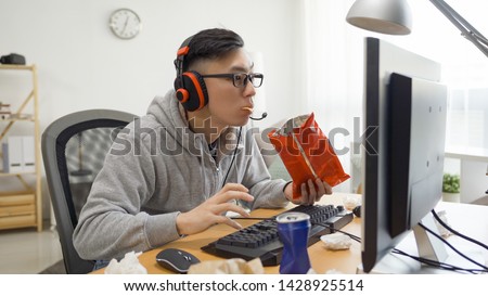 Teenager boy high school student looks at the computer screen holds chips in hand and plays video games on weekends. asian guy in grey sweatshirt eating snack and surfing internet watch anime cartoon