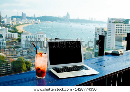 Laptop and glass of summer cocktail on wooden table, on bright beautiful panoramic background of Pattaya city, Thailand