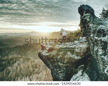 Hiker and photo enthusiast stay with tripod on cliff. Peak with woman taking photos in morning sunrise