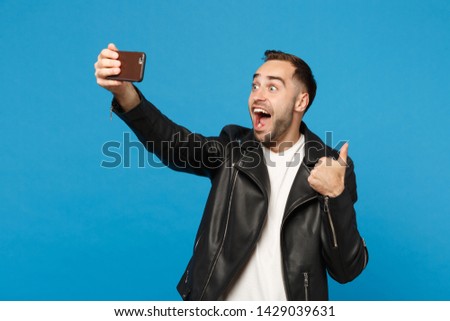 Stylish young unshaven man in black jacket white t-shirt oing selfie shot on mobile phone isolated on blue background studio portrait. People sincere emotions lifestyle concept Mock up copy space