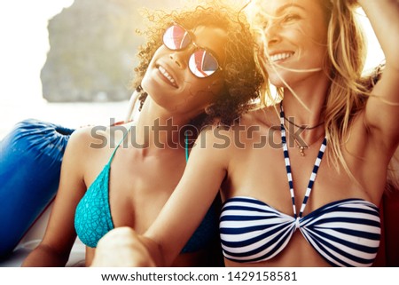 Two smiling young female friends wearing bikinis sitting together on a boat enjoying a sunny day out on the open ocean