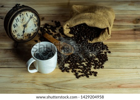 A white coffee mug with smoke and brown roasted coffee beans in wooden spoon and spilled from sackcloth on pine wood floor with old vintage alarm clock background.