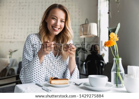 Beautiful young blonde woman sitting at the cafe indoors, eating cake, taking photo with mobile phone