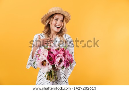 Portrait of a lovely young blonde girl wearing summer dress standing isolated over yellow background, holding peonies bouquet