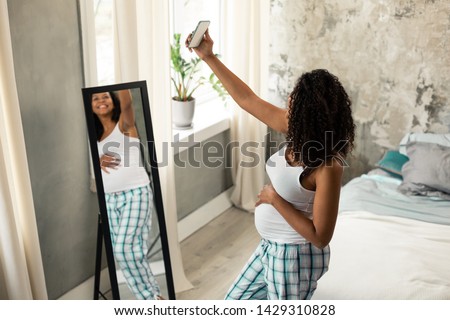 Taking a picture. Happy smiling pregnant woman standing in front of her mirror at home and taking a selfie.