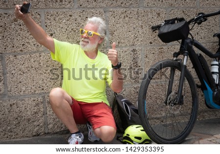 Elderly man with white hair and beard, raises his thumb in a sign of victory. He takes a selfie with his cell phone near his bicycle. Brick wall as a background. Colorful way, with a yellow helmet