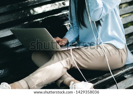 Young brunette girl sits on a bench in a city park, works on a notebook and listens to music on headphones
