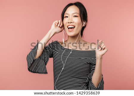 Photo of beautiful asian woman dressed in basic wear singing and listening to music with earphones isolated over red background in studio