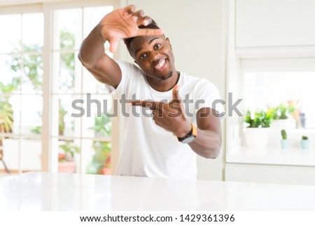 Handsome african american man on white table at home smiling making frame with hands and fingers with happy face. Creativity and photography concept.