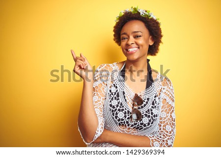 Young african american woman with afro hair wearing flowers crown over yellow isolated background with a big smile on face, pointing with hand and finger to the side looking at the camera.