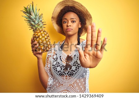 Young african american woman with afro hair holding pineaple over yellow isolated background with open hand doing stop sign with serious and confident expression, defense gesture