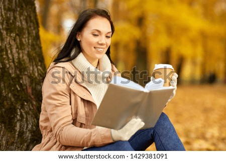 season, technology and people concept - young woman reading book and drinking takeaway coffee from paper cup in autumn park