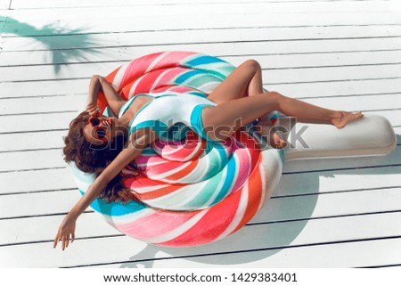 Slim girl having fun on colorful Inflatable lollipop pool float. Bikini model in bright swimwear sun tanning on tropical bech at summer day. 