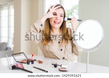 Beautiful young woman using make up cosmetics applying color using brush with happy face smiling doing ok sign with hand on eye looking through fingers