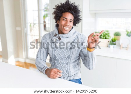 African American man wearing winter sweater with a big smile on face, pointing with hand and finger to the side looking at the camera.