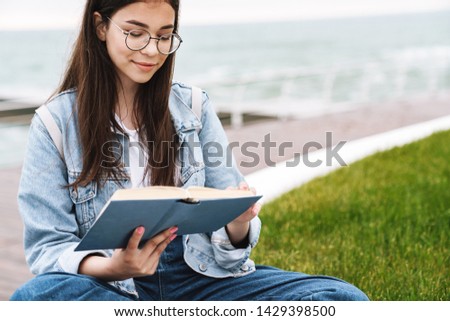 Image of thoughtful lovely teenage girl wearing eyeglasses reading book while sitting on green grass by seaside