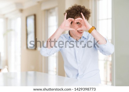 Young business man with curly read head Doing heart shape with hand and fingers smiling looking through sign