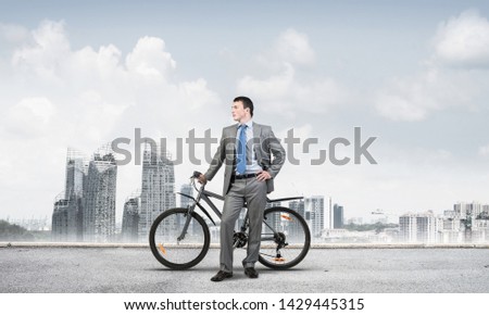 Young man wearing business suit and tie standing on asphalt road with bike. Businessman with bicycle on background of blue sky above megalopolis. Male cyclist holding bicycle, having break in riding.