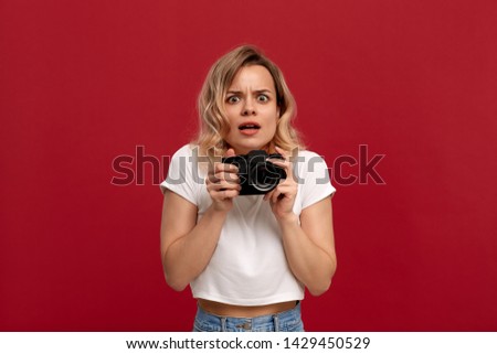 Portrait of a girl with curly blond hair dressed in a white t-shirt standing on a red background. Model with shocked emotions holds retro film camera. 