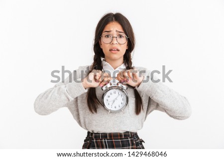 Photo closeup of disturbed teenage girl wearing eyeglasses frowning while holding alarm clock in hands isolated over white background